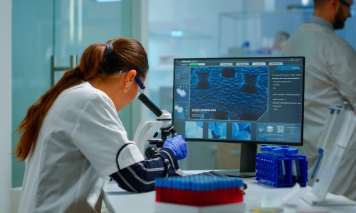 Biotechnology scientist researching in laboratory using microscope and typing on pc. Chemist examining virus evolution using high tech for scientific research of vaccine development against covid19